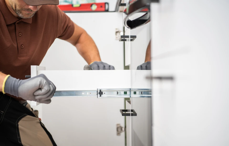 Un homme en combinaison bleue installe ou répare des éléments de cuisine, utilisant un marteau pour ajuster des charnières de placard